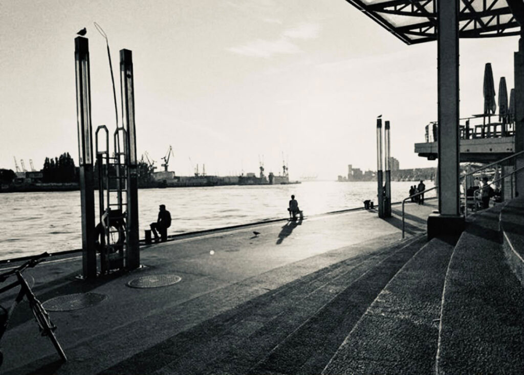 Schwarz-Weiß-Fotografie: Anleger am Hamburger Hafen im Gegenlicht, eine Person sitzt am Elbufer, im Hintergrund Werftkräne